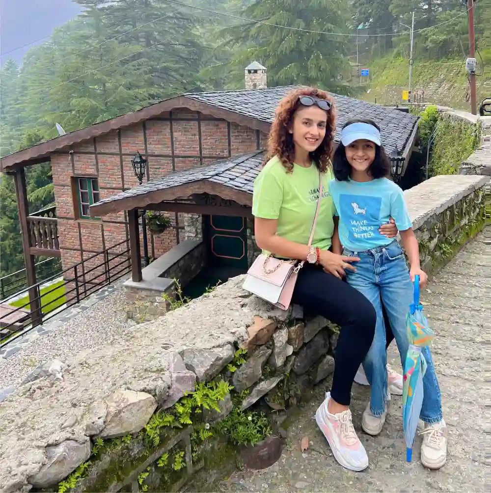 Two people sitting on a stone wall in front of a rustic building with greenery in the background