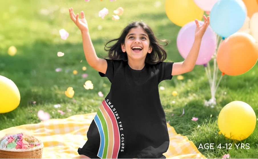 Child in a black t-shirt with colorful text, standing outdoors with balloons and picnic blanket.