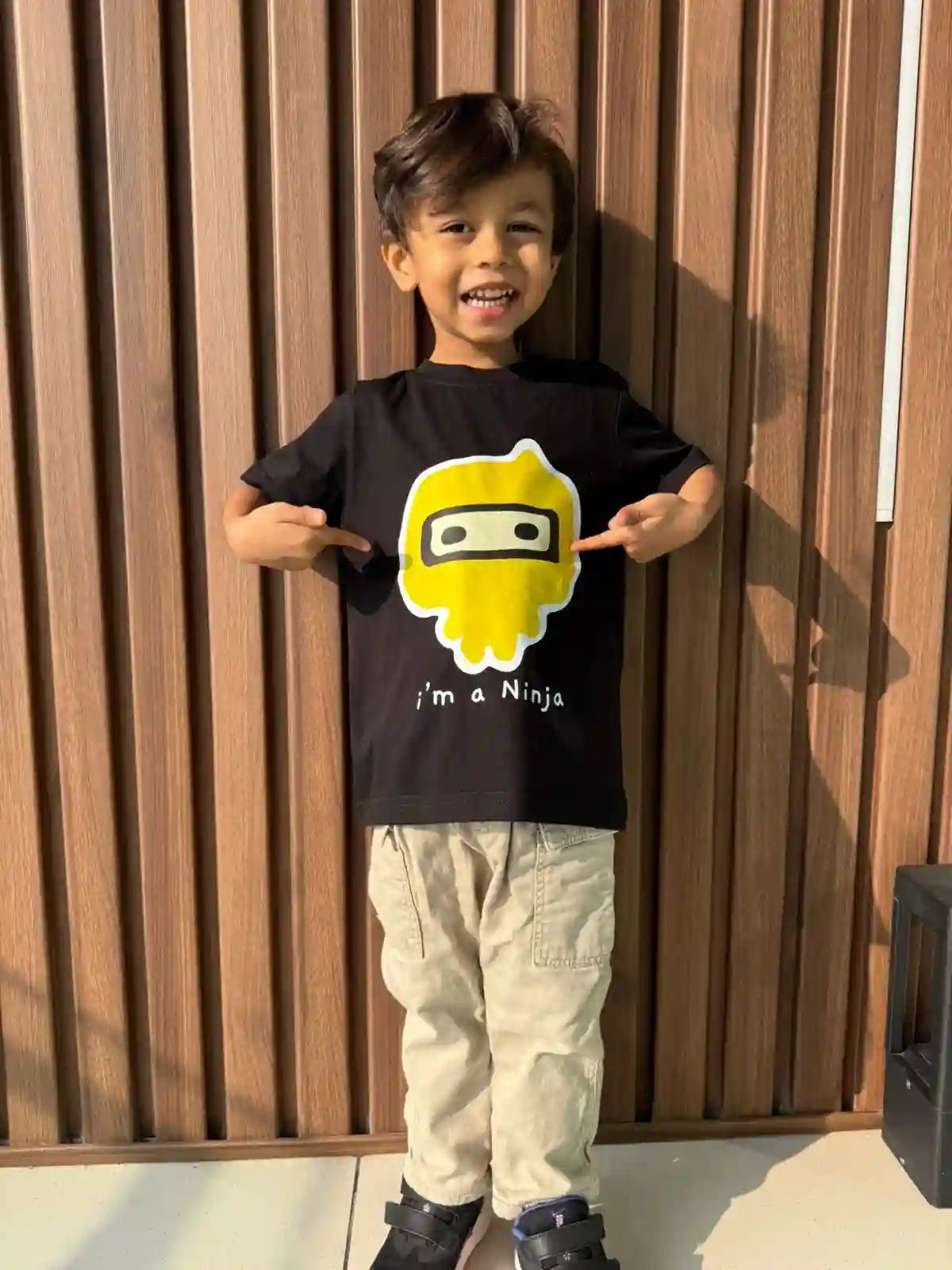 Child wearing a black t-shirt with a yellow graphic and text, standing against a wooden wall.