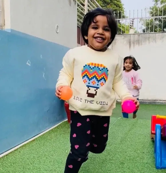 Child holding colorful balls in an outdoor setting with a blue wall and green ground.